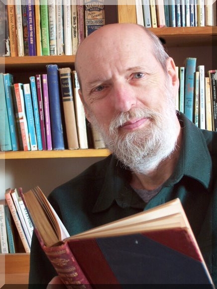 Photo. A man holding an open book while sitting in front of a bookshelf.